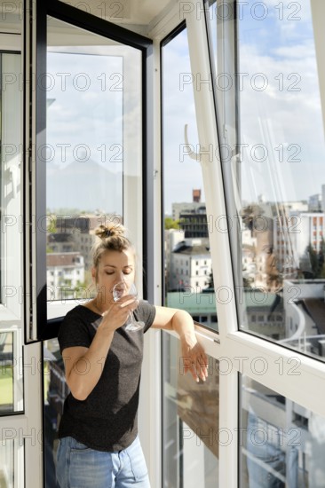 A woman leans against a balcony railing, sipping a drink and looking out at the vibrant cityscape. Sunlight pours in through large windows, illuminating her relaxed posture and quiet moment