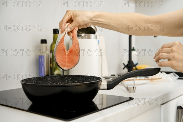 A hand is lifting a fresh salmon fillet above a black frying pan on a stovetop. The kitchen appears bright and modern, with condiments and utensils visible in the background