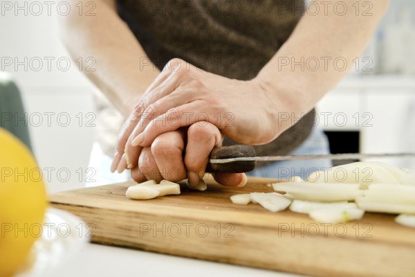 Unrecognizable woman crush garlic to finely chop it for sauce on a wooden cutting board in a bright kitchen