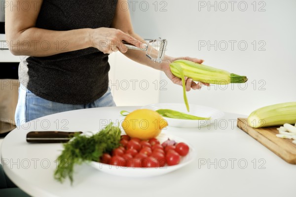 A person prepares fresh vegetables in a modern kitchen, peeling zucchini while other ingredients, including cherry tomatoes and lemon, are arranged neatly on the table