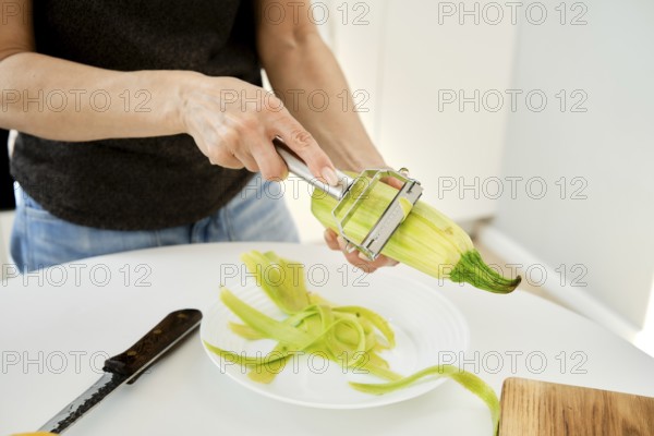 A person is peeling a zucchini in a bright kitchen. Fresh zucchini is held in one hand while a peeler removes the skin