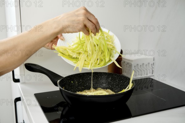 A hand pours freshly cut zucchini noodles into a sizzling pan on an electric stovetop. The kitchen is bright and modern, showcasing meal preparation for an evening meal