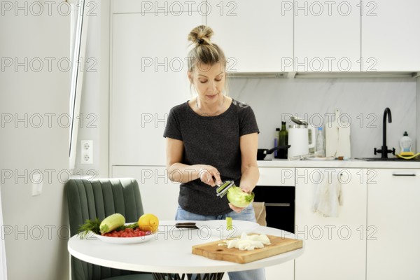 A middle-aged woman is skillfully peeling a green zucchini at a kitchen table