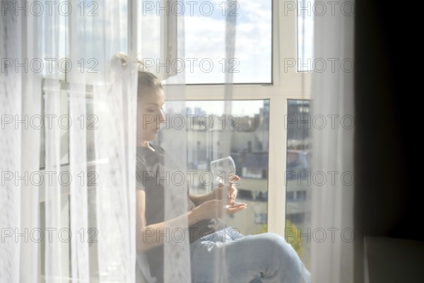 Sunlight streams through sheer curtains as a woman sits peacefully by the window. She gazes thoughtfully at a glass, enjoying the tranquil moment while overlooking a bustling city