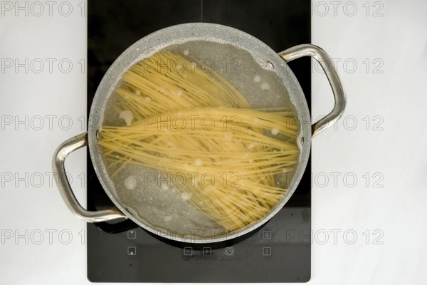 Long strands of pasta are submerged in boiling water inside a pot on an induction stovetop. The kitchen environment is clean and modern, showcasing the cooking process clearly