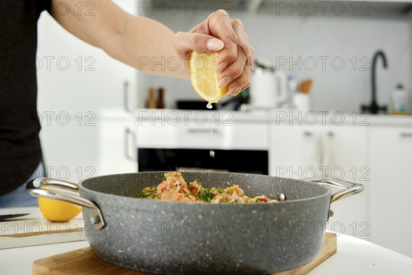 A person is preparing a seafood dish by squeezing fresh lemon juice into a skillet. The modern kitchen features sleek appliances and a vibrant atmosphere, enhancing the cooking experience