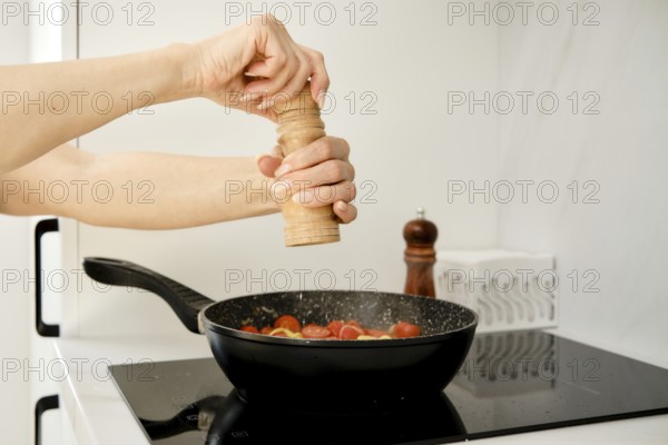 A hand is grinding pepper over a skillet filled with fresh tomatoes. The setting is a modern kitchen featuring a sleek countertop and cooking utensils, creating a warm cooking atmosphere