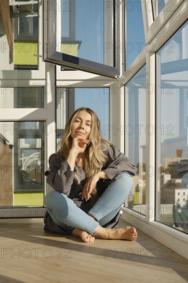 A middle-aged woman sits comfortably on a wooden floor of balcony in a bright modern apartment. She enjoys the sunlight streaming through expansive windows, surrounded by a vibrant cityscape