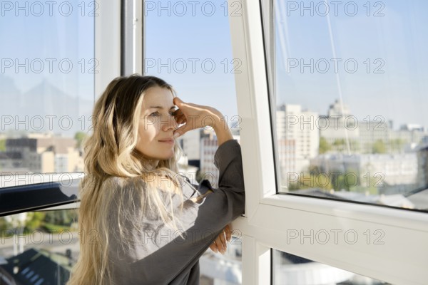 A woman stands by a wide window, thoughtfully looking out at a bright city landscape. Her long hair cascades around her shoulders as sunlight streams into the room, creating a serene atmosphere