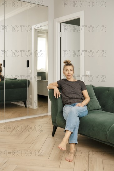 A woman sits comfortably on a green couch in a modern living room. She poses casually, with her bare feet resting on the floor. Natural light fills the space, highlighting the minimalist decor