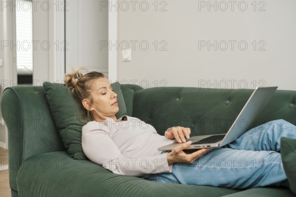 A woman with a relaxed demeanor is laying on a green couch in a bright living room, focused on her laptop