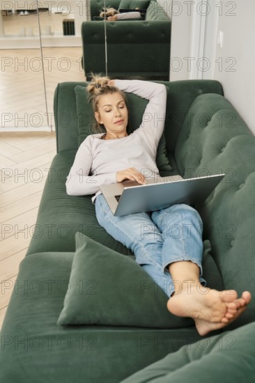 Middle-aged woman is lounging on a green couch, scrolling on her laptop in online shop choosing goods