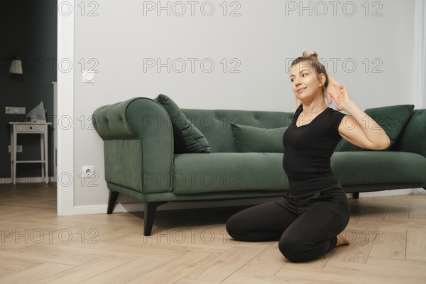 A middle-aged woman in a black outfit is kneeling on the wooden floor, practicing yoga in a stylish living room with a green sofa