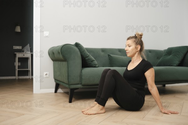 A woman dressed in comfortable workout attire is seated on the floor in a living room. She engages in a fitness routine, focusing on her posture and breathing
