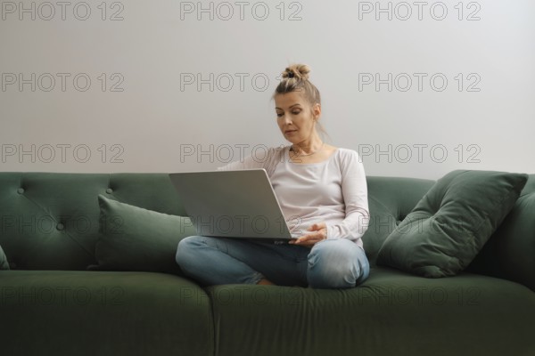 A woman sits cross-legged on a soft green couch, focused on her laptop. Natural light fills the relaxing living room, creating an inviting atmosphere for work or leisure in the afternoon