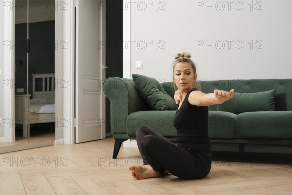 A middle-aged woman practices stretching exercises on a wooden floor in a cozy living room. The space features a green couch and a mirror, providing a calming atmosphere for her yoga routine