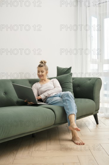 A woman enjoys her time on a green couch, focused on her laptop. Sunlight fills the bright living room, where she appears relaxed and engaged in her work, dressed casually in comfortable clothing