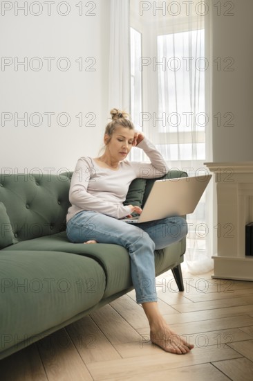 A woman is focused on her laptop while seated on a soft green sofa. She appears thoughtful, with her hand resting on her forehead