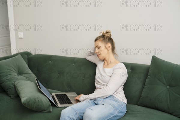 Middle-aged woman is seated on a green couch in a well-lit living room, working intently on her laptop. She appears focused and concentrated, dressed comfortably in casual clothes
