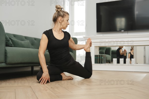 A middle-aged woman in athletic wear performs leg stretching on the floor at home, combining flexibility and balance