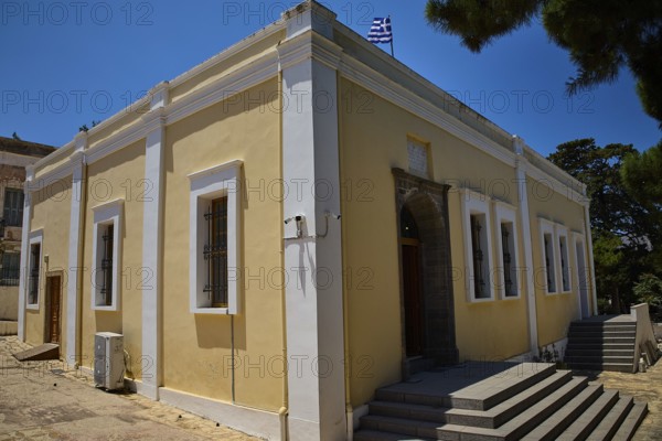 Yellow building with flag, surrounded by trees. Historic architecture with white window frames, Archaeological Museum, Agia Marina, Leros, Dodecanese, Greek Islands, Greece