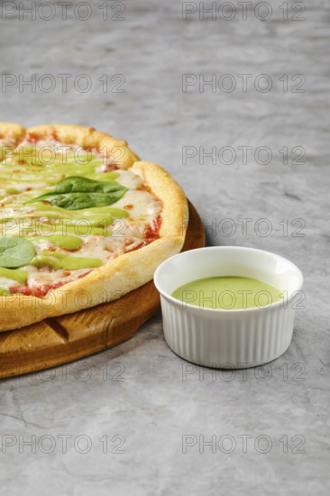 This dish features a freshly baked pizza with a golden crust adorned with toppings like fresh greens and a flavorful tomato base. A small bowl of vibrant green sauce complements the pizza perfectly