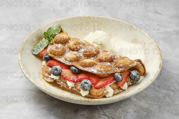 Colorful bubble waffles are served with fresh strawberries and blueberries, topped with creamy filling and a scoop of ice cream. The dish is garnished with mint leaves for a refreshing touch