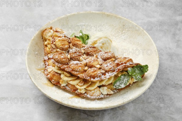 A beautifully presented dessert showcases a crispy waffle filled with sliced bananas and caramel, topped with powdered sugar and a scoop of vanilla ice cream. Mint leaves add a pop of color