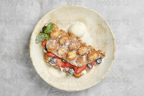 A golden bubble waffle is plated with strawberries, blueberries, and a scoop of vanilla ice cream. Fresh mint leaves add a touch of color, creating a delightful dessert ready to be enjoyed
