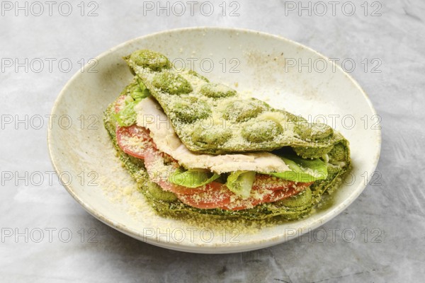 A vibrant sandwich made with green dough stacked with fresh slices of chicken, tomatoes, and lettuce. The dish is topped with grated cheese, presented in a rustic bowl suitable for casual dining