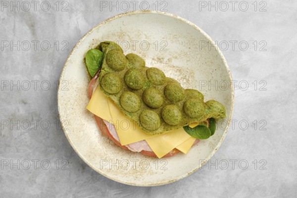 A creatively prepared sandwich features a vibrant green, fluffy waffle. Inside, layers of ham and cheese are complemented by fresh greens