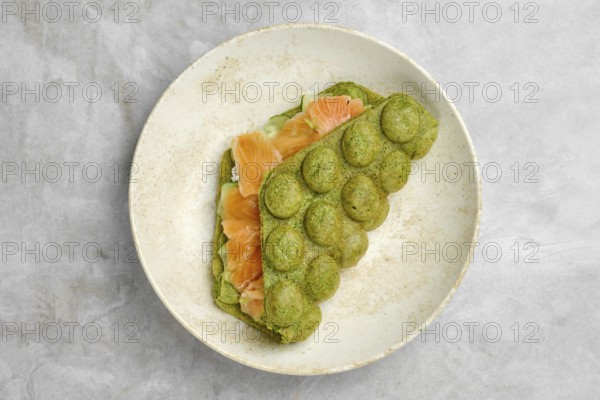 A unique green spinach waffle is folded over slices of smoked salmon, carefully arranged on a simple white plate. The dish showcases a blend of flavors and textures, perfect for a gourmet breakfast