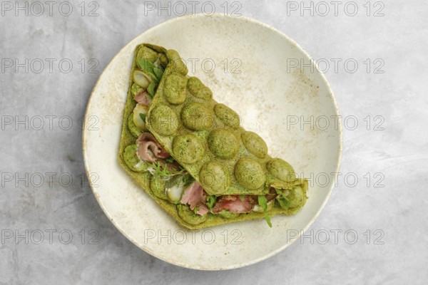 A green wrap made with spinach or herbs contains layers of fresh vegetables and meat, resting on a simple plate. This dish is presented in a relaxed cafe setting, perfect for a light meal
