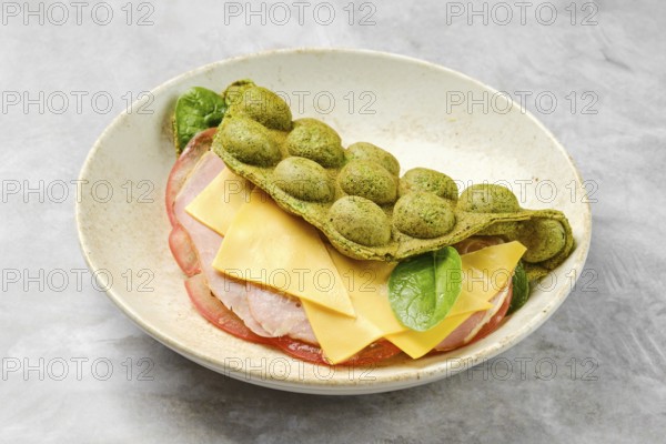 A unique green wrap made from a blend of ingredients encases layers of ham, cheese and spinach waffle, resting elegantly in a stylish bowl. The dish showcases vibrant colors and textures