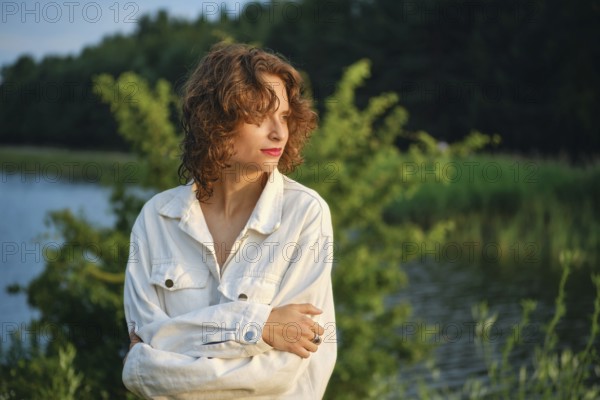 On a hot day, a woman stands near the calm lake, basking in the sunlight. Surrounded by lush greenery, she admires the serene water, lost in thought and enjoying the warmth of summer