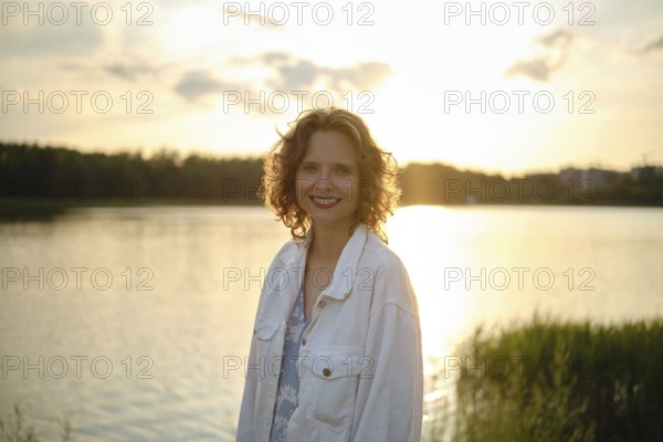 A woman stands near a shimmering lake on a hot evening, her curly hair catching the sunlight. The warm tones of sunset reflect on the water, surrounded by lush greenery and a peaceful atmosphere