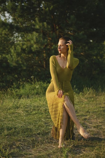 On a warm day, a woman sits gracefully by the lake surrounded by lush greenery. Wearing a fishnet knitted dress, she enjoys the tranquility of nature and the refreshing atmosphere of summer