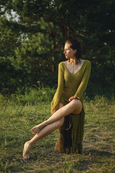 A woman enjoys a hot day by the lake, sitting gracefully on a rustic log. The sun casts a warm glow on her, highlighting her relaxed pose amidst lush greenery