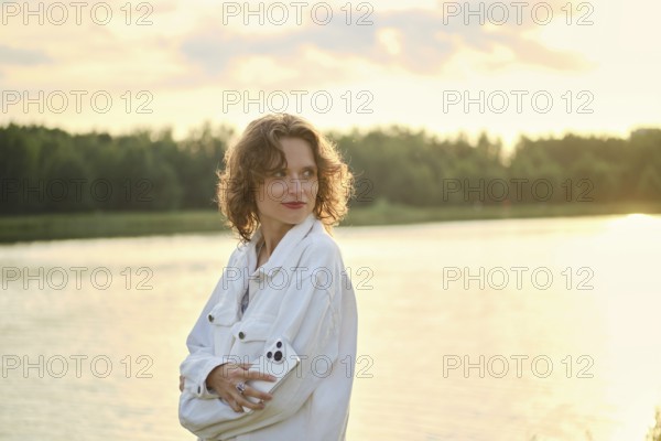 Cute woman with curly hair stands by the lake at sunset. She look back while holding smartphone in her hand