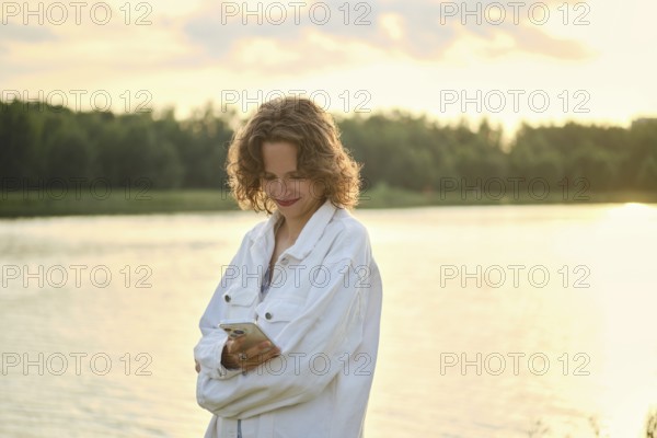 A person stands by the lake on a hot day, enjoying the sunny weather. The golden light creates a serene atmosphere as she glance at her phone, reflecting a moment of relaxation by the water