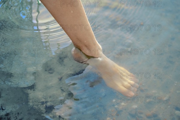 One foot dips into the refreshing lake water, creating gentle ripples on the surface. The bright sun shines down, making the day perfect for enjoyable outdoor activities and relaxation by the water