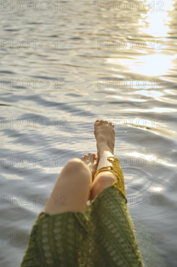 Enjoying a hot day at the lake, woman relaxes with her feet dipped in the calming water. The sun creates sparkling reflections, enhancing the tranquil atmosphere of the moment