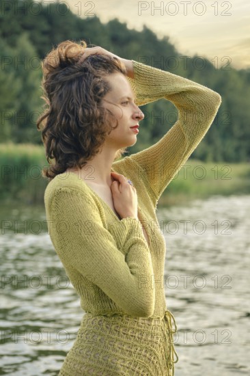 On a hot day, a woman stands gracefully by a serene lake touching her curly hair. She seems lost in thought while embracing the beauty of her natural surroundings