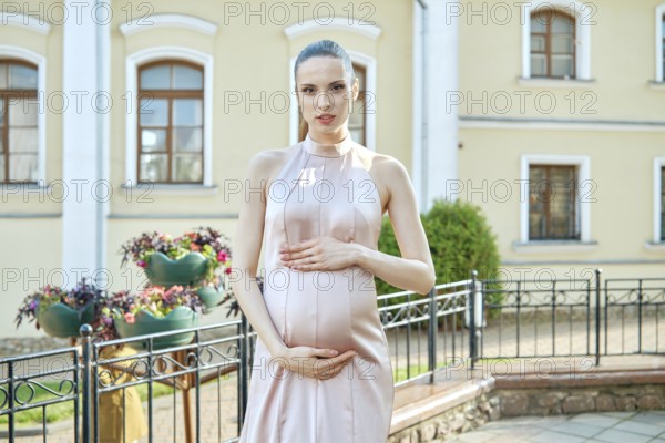 A woman in a flowy dress showcases her pregnancy in a charming outdoor setting. Surrounded by a beautiful garden, she holds her belly with a joyful expression
