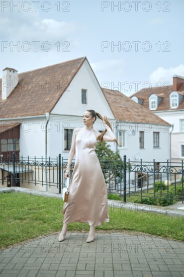 A stylish woman is posing gracefully in a lovely courtyard, dressed in a flowing pink dress. The background features charming architecture and lush greenery under a clear blue sky