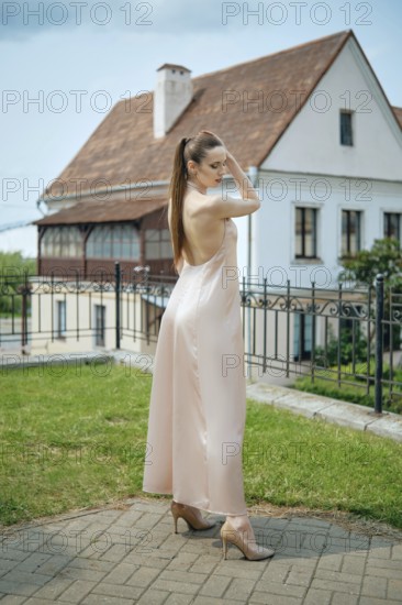 A young woman showcases her stylish outfit in a soft-colored dress while standing in a grassy area. Behind her is a charming vintage house with classic architecture, all under a bright sky