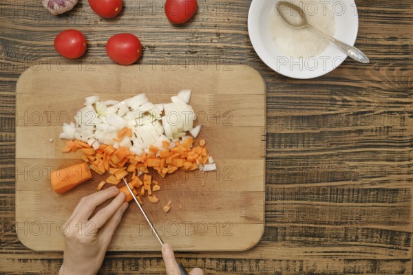 A person cutting diced carrots and onions on a wooden cutting board. Fresh tomatoes and garlic can be seen in the background, showcasing a vibrant kitchen environment