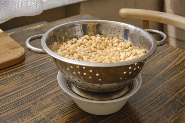A colander filled with soaked white beans is resting above a bowl to catch excess water. Natural light filters into the cozy kitchen, highlighting the rustic wooden surface and homey atmosphere