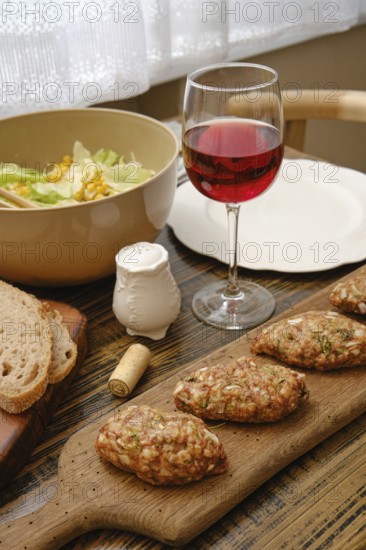 A table is set with a wooden board of homemade patties, a bowl of fresh salad, a glass of red wine, and slices of bread. Sunlight streams through the window, enhancing the cozy atmosphere