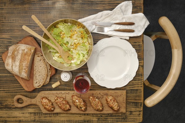 A vibrant salad with greens and corn in a bowl next to a loaf of rustic bread. Savory meat patties are on a wooden board, accompanied by a glass of wine, creating a wholesome lunch setting